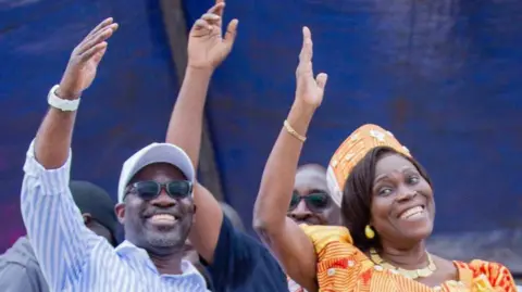 MGC Simone Gbagbo dressed in orange stands to the right of  Charles Blé Goudé  in a stripped blue and white shirt in glasses