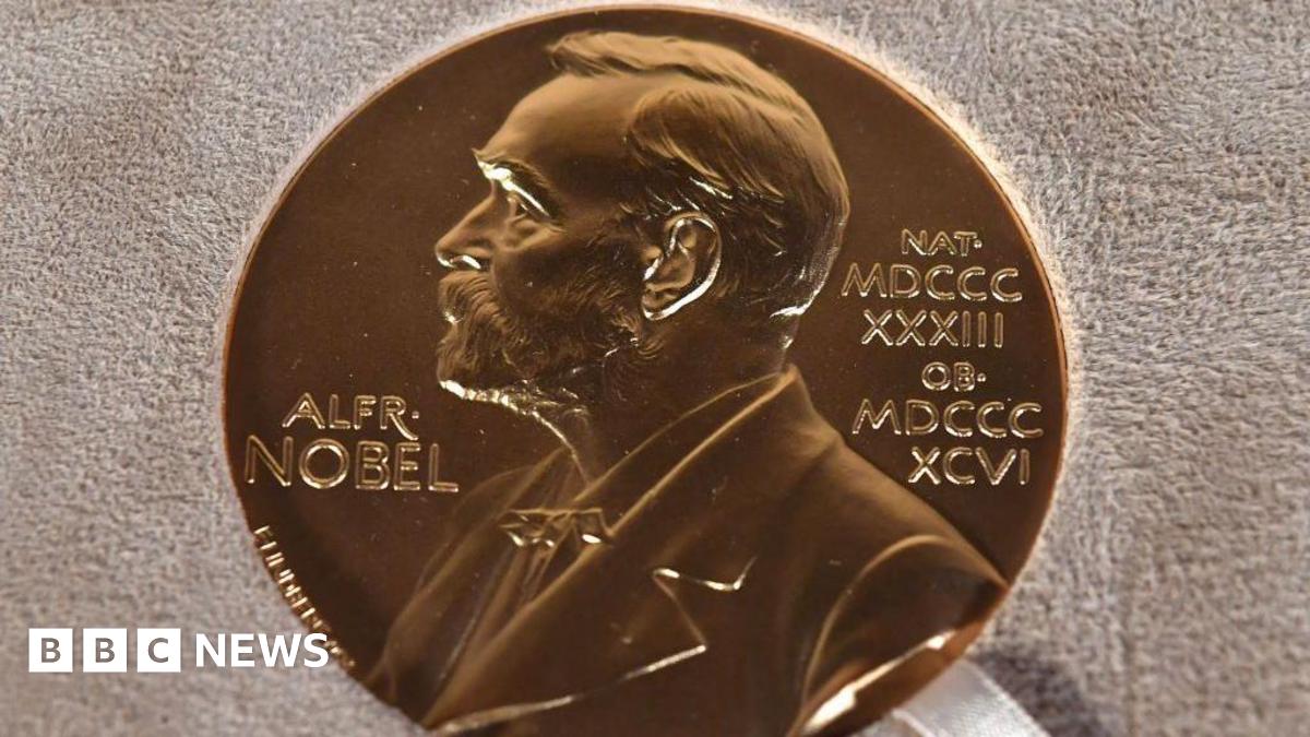 Kemi Badenoch wearing a white jacket and white t-shirt is pictured waving to the camera. There is a union jack pictured to the left.