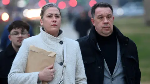 Reuters Caroline Darian, a woman with her blonde hair pulled back and a white herringbone wool coat, clutching a manila envelope, and David Pelicot, a man with dark hair and a dark wool coat over a mid-grey suit, walk outside the courthouse in Avignon in December last year, with a determined expression on their faces