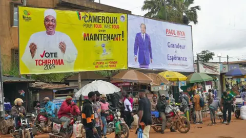 Michel Mvondo / BBC People hang around on motorbikes on a dustry road where two campaign posters have been erected - one for Biya and the other for Tchiroma.