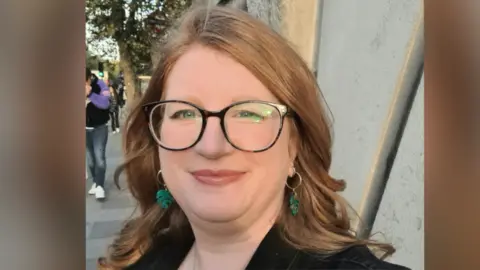 Headway Norfolk and Waveney Claire Boothby-Barnbrook a woman who is standing outside, she has curled hair and is looking directly at the camera and smiling. She is wearing a black outfit with black frame glasses and has green leaf shaped earrings.
