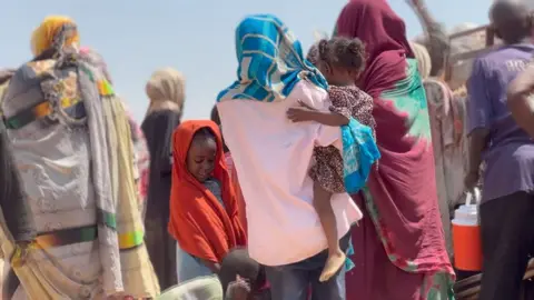 A woman, in a pink shirt and blue scarf, with her back to the camera holds a child on her hip. Another child can be seen on her left.