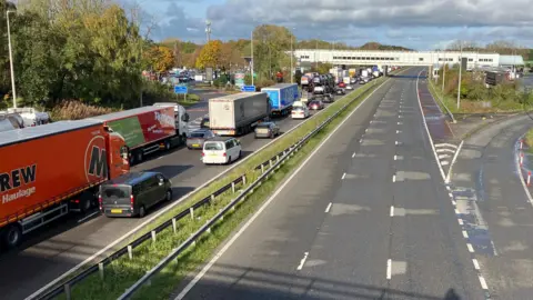 Ollie Samuels/BBC A long line of cars, vans and lorries queues on one carriageway of the M6, under the bridge at Charnock Richard services, while the other carriageway is completely empty