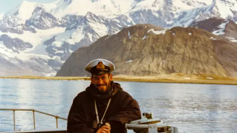Roly Woods Commander Woods standing on a ship smiling with mountains behind him wearing a navy cap
