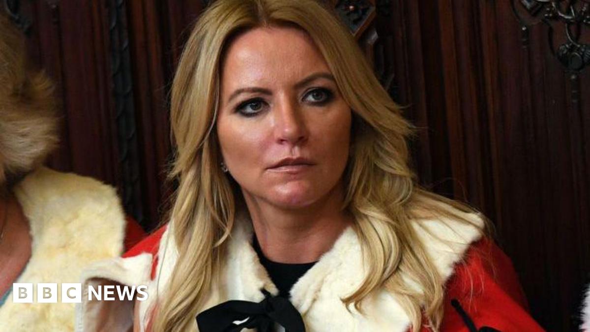 Michelle Mone in her House of Lords robes. She has long blonde hair and is looking off camera, sitting on the red benches wearing white ermine and red robes