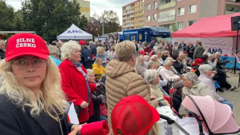 A crowd of people surrounding a set of marquees, many of whom are wearing the Silné Česko/Strong Czechia red hats including a blonde woman in the foreground. The attendants are larger older people. 
