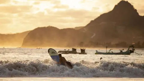 Getty Images Back view of a man lying on his surf board at sunset as a wave crashes into him. In the background are fishing boats and a large hill.