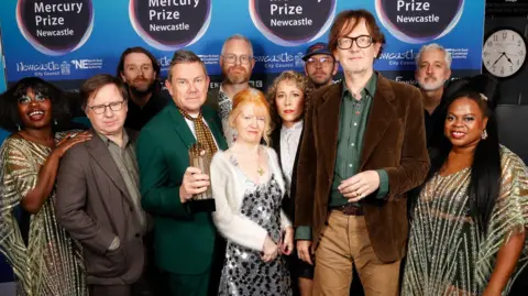 Getty Images The band Pulp and accompanying musicians smiling into the camera. There are 11 people dressed in colourful clothing. Jarvis Cocker is standing at the front, second from the right and has brown, sweepy hair and is wearing large glasses. He is wearing a brown cord blazer and a green shirt underneath. He is wearing lighter brown trousers. They are standing in front of a blue Mercury Prize board.