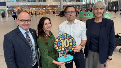 Mark Adair has receding dark hair. He is wearing glasses, a blue shirt and jacket and bright patterned tie. Julie Reid has long auburn hair and is wearing a green dress. She is holding the Book Week symbol, which is in the shape of a tree with multi-coloured leaves and the word 10th in the centre. Conor McKee has brown hair, parted to one side. He is wearing glasses and a white shirt. Jacqueline Kennedy has shoulder length fair hair and is wearing a dark blouse and jacket. They are standing on the concoures of a bus station