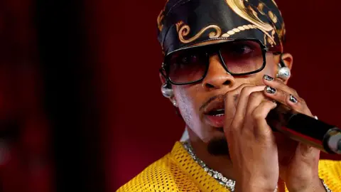 Getty Images D4vd sings into a microphone. He wears a yellow sports jersey, lots of diamond jewelry, a silk scarf tied around his head and sunglasses