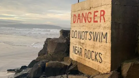 BBC Portstewart Strand. A large sign reads: 'Danger – Do Not Swim Near Rocks.' It is positioned beside a wide area of jagged black rocks, with the shoreline visible in the background.