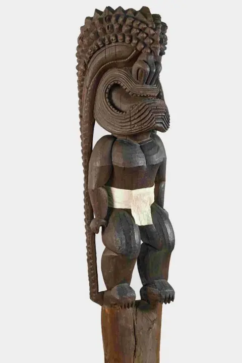 Trustees of the British Museum A wooden sculpture of a human-like figure wearing a large helmet that covers the face and extends at the back to the floor. Is it wearing a white loin cloth. 