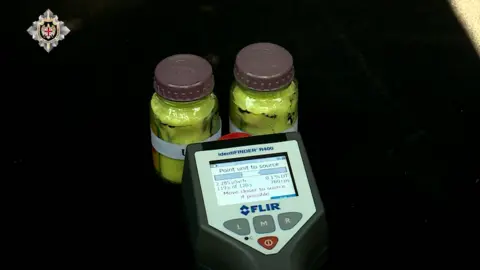 SSG Two glass jars containing a yellow substance in a car boot with a dosimeter pointing at them.