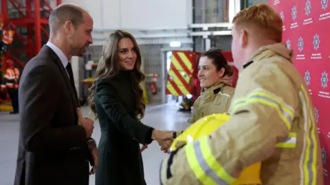 Chris Jackson/PA Wire A male firefighter with short ginger hair shakes hands with Catherine, Princess of Wales while a young female firefighter with dark hair stands beside them, smiling.  Prince William is standing beside his wife, talking to the recruits.  The firefighers are wearing beige safety suits and carrying bright yellow safety helmets. There is a red fire safety vehicle parked behind them.