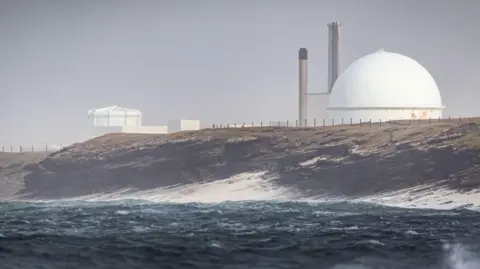 Getty Images The power station is a complex of buildings and structures, including metal sheds with peaked roofs, a chimney and a large sphere. It is situated on a rugged coastline. The sea is dark blue and choppy.