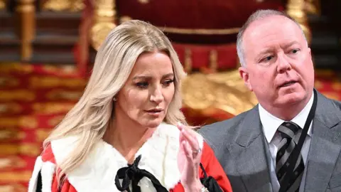 Getty Images Baroness Mone and her husband both stand on an ornate red and gold carpet in a richly decorated room. She wears a ceremonial red robe with white fur trim and gold detailing. Her husband is dressed in a grey suit with a black tie.