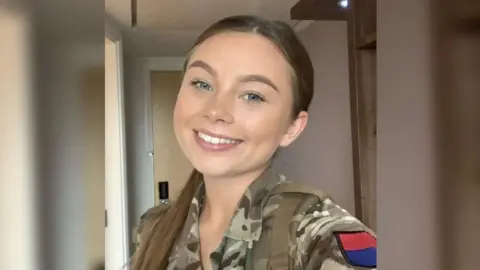 Family photo A young woman wearing army camouflage is smiling in a selfie style photo.  She has her hair pulled back into a ponytail, which is sat over her shoulder. There is a red and blue patch on the top of her sleeve.  
