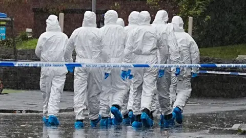 PA Media A group of forensic investigators in white PPE and blue gloves and shoe coverings, walk away from the camera behind a line of blue and white police tape. 