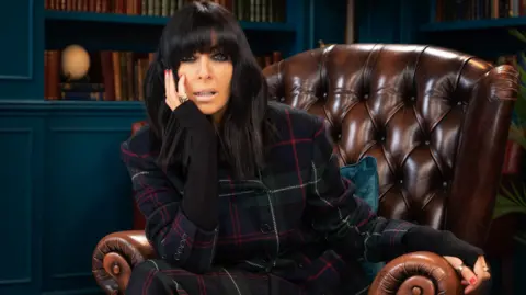Claudia Winkleman sitting in an armchair in the Traitors castle in Scotland