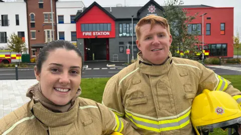 Caoimhe McNeice and Piarais McCaffrey posing for a photo in front of the fire station training facility outside Cookstown.  Caoimhe has dark hair tied back in a ponytail or bun and Piarais has short ginger hair.  They are wearing beige protective suits with reflective stripes. There is a red and black fire station behind them and tall buildings to the left. 