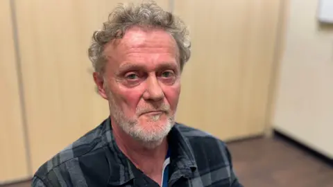 A man with curly grey hair is looking into the camera. He has a short white beard and is wearing a grey check button up shirt. 