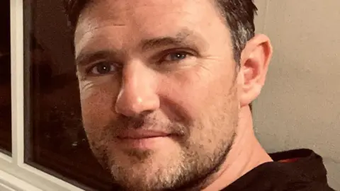 Dan Henshaw Dan Henshaw in a selfie. He has brown greying hair and a patchy beard. He is a white man. He looks to be in a living room or somewhere at home.