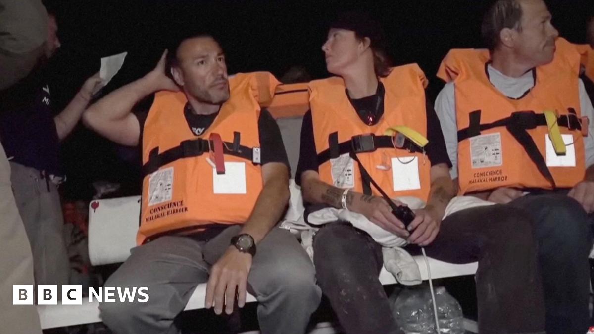 Three crew members on flotilla vessel Conscience - two men and a woman in the middle - are seen sitting on a white bench and wearing orange safety vests and dark trousers