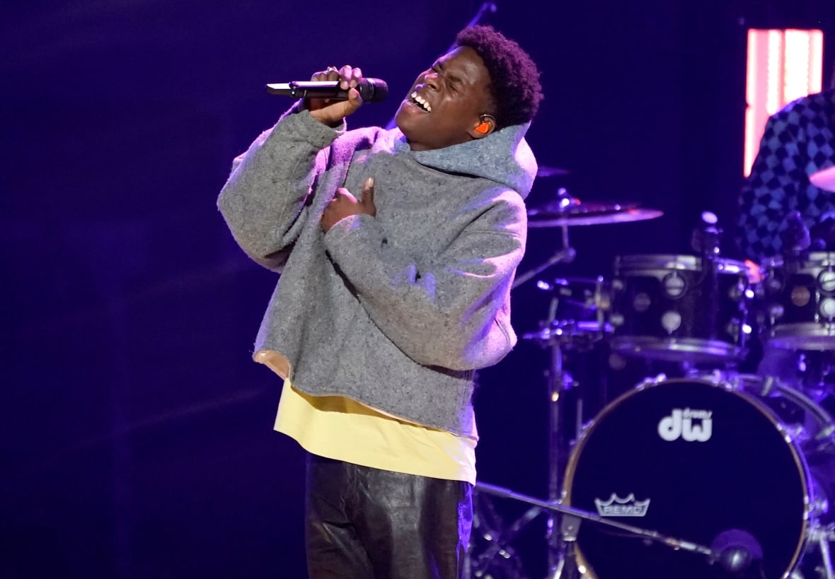 A man holds a mic and sings on stage. He is wearing a grey hoodie.