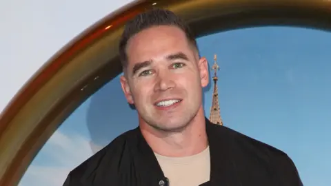 Getty Images Kieran Hayler smiling at the camera. He has short, dark hair and is wearing a beige shirt under a black jacket.