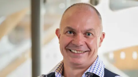 Prof David Strain University of Exeter's Prof David Strain smiles broadly. He is wearing a blue and white chequered shirt and a grey waistcoat.