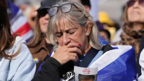 PA Media A woman with blonde hair, draped in a blue and white Israeli flag, closes her eyes and bows her head, with one hand held over her mouth. In the other, she holds a flyer displaying a picture of a male Israeli hostage, along with his name - David Cunio - and age - 34 - printed in red, capital letters, in both English and Israeli. Other attendees are pictured out of focus in the background.