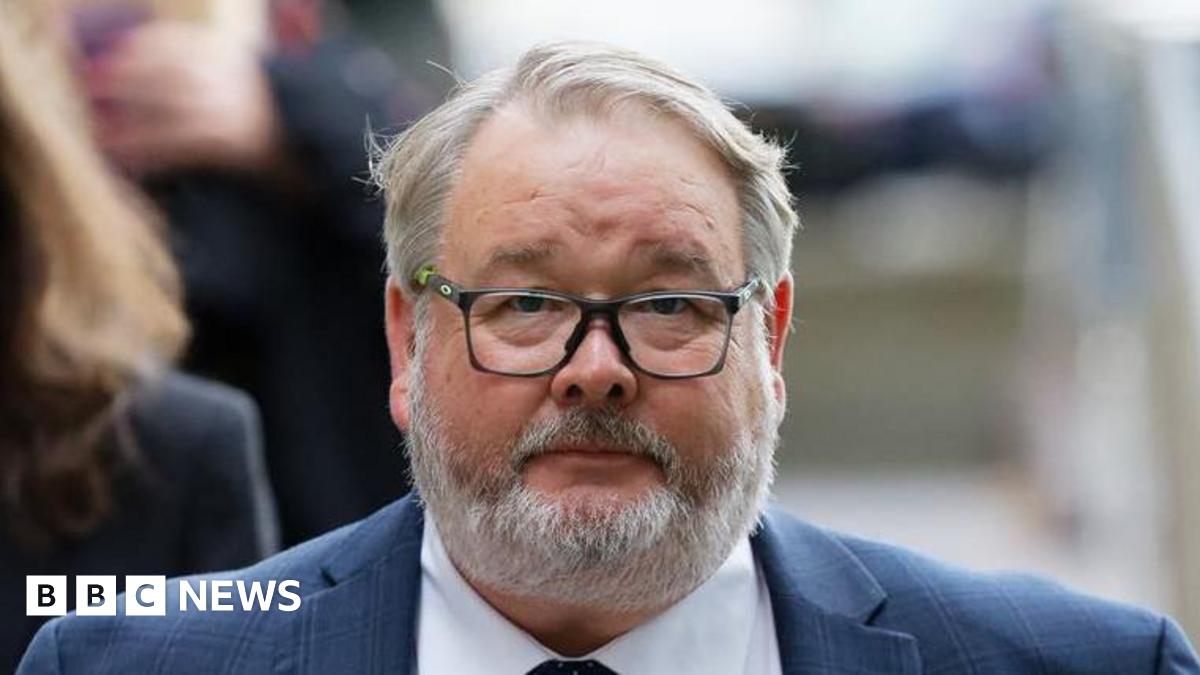 Close up of Christopher Mills outside court. He wears a blue suit, a white shirt and has black glasses. He has short grey hair and a grey beard.