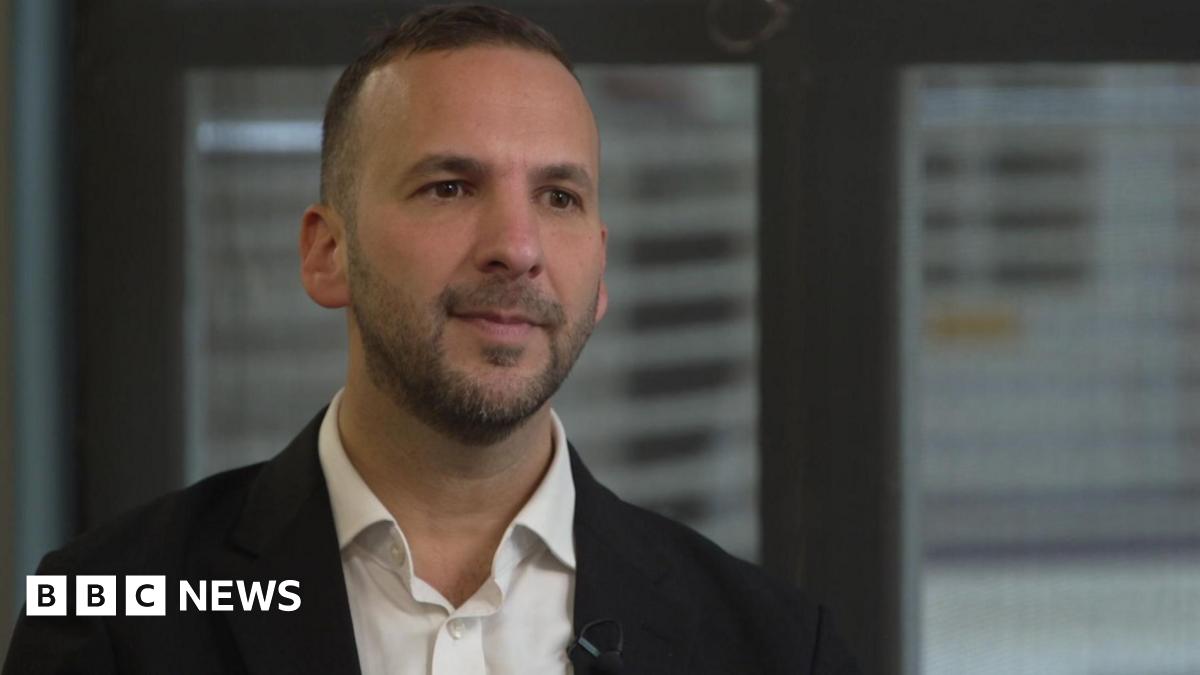 Zack Polanski in a dark jacket and white shirt sitting in front of two office windows.