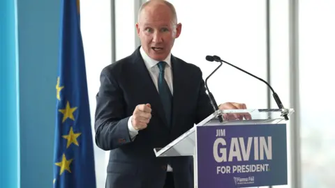 PA Jim Gavin has receding hair. He is wearing  a dark suit and a green tie and pointing towards the camera.