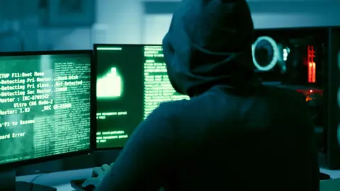 Getty Images A person wearing a black hoodie sits at a computer with multiple screens running green code.