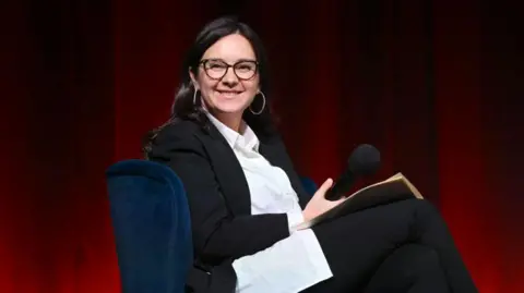Getty Images Bari Weiss in a black blazer and white shirt with big hoop earrings and glasses, holds a microphone in her hand