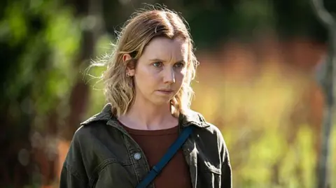 BBC/The Ridge Production Ltd A young blonde woman looks somewhat distressed and dishevelled, standing outside with greenery behind her. She has a serious look, staring off to the left and wears a brown T-shirt, dark denim jacket and a cross-body bag.