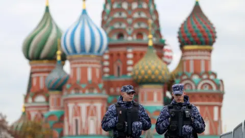 Reuters Two men in blue and black camouflaged uniforms and hats and black vests talk as they walk in front of a red stone cathedral with blue, green, red and gold designs on its domes.