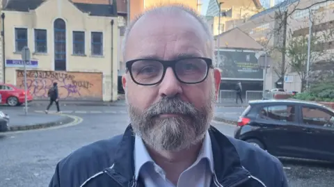 BBC A man with short balding grey hair and a grey beard, looking at the camera. He is wearing glasses, a navy jacket and a blue shirt.