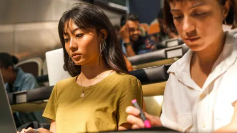 Getty Images Students sit in a bright lecture hall with tiered seating. Two female students are in focus. The one of the left wears a mustard-yellow T-shirt, a gold necklace, has long dark hair with a fringe, and large hoop earrings. She is typing on a laptop. The student on the right, in a white button-up short-sleeved shirt with short dark hair, holds a pink highlighter over notes.