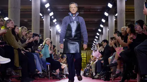 Getty Images Actor Richard E Grant in a black apron, blue jumper and navy trousers walking the runway for Miu Miu.