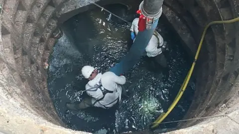 PA Media Two workers in protective suits and helmets standing in in murky water inside a large sewer chamber.