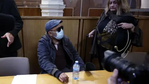 GUILLAUME HORCAJUELO/EPA/Shutterstock A man in a cap and anorak and wearing a mask sits at a desk while surrounded by photographers