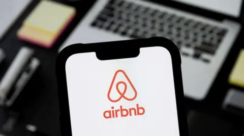 Getty Images A phone with the Airbnb app is depicted in the foreground, and laptop is pictured in the background