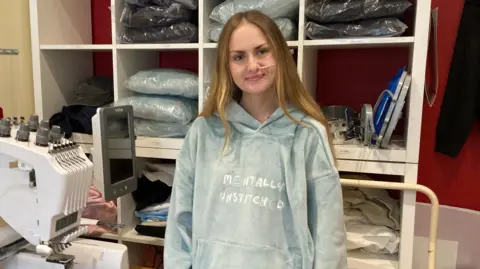 BBC Millie Hayward stands next to an industrial embroidery machine in her home studio. She wears a greyish blue hoodie with the words "mentally unstitched" embroidered on and stands in front of boxed shelves of packaged hoodies. She has long blonde hair and a medical nasal tube.