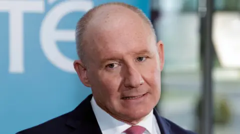 PA Media A close-up of Jim Gavin. He is an older man, with balding/short grey hair. He is mid-speech and is standing in front of a large blue and white RTÉ board. He is wearing a dark suit jacket, a white collared shirt and a pink tie.