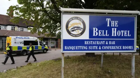PA Media A large blue and white sign in the grounds outside The Bell Hotel in Epping. In the background there are two police officers in hi-vis jackets and a police van.