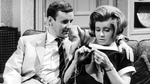 Richard Briers & Prunella Scales in Marriage Lines. Briers has his arm around Scales, who is shown knitting.