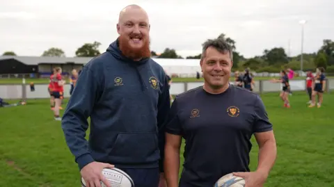 James Phipps and Gavin Vickers are both wearing navy and gold Worcester kit and holding rugby balls.  James is tall with a ginger beard, Gavin is shorter with brown hair. 