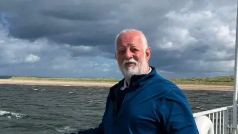 SUSAN JOHNSON A photo of Barrie, with white hair and stubble, he wears a blue jumper and smiles while being photographed in front of the sea.
